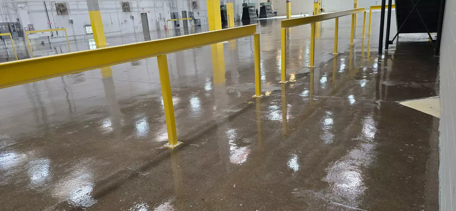 A wide view of a large commercial floor with yellow safety railings and a wet, reflective concrete surface during the sanding phase.