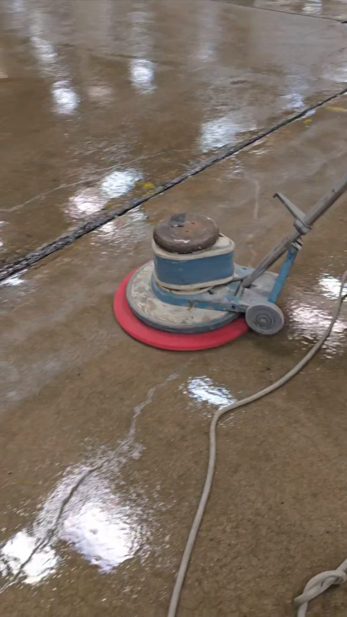 A rotary floor machine with a round red pad working across a wet concrete floor during the sanding phase, with its power cord trailing behind.