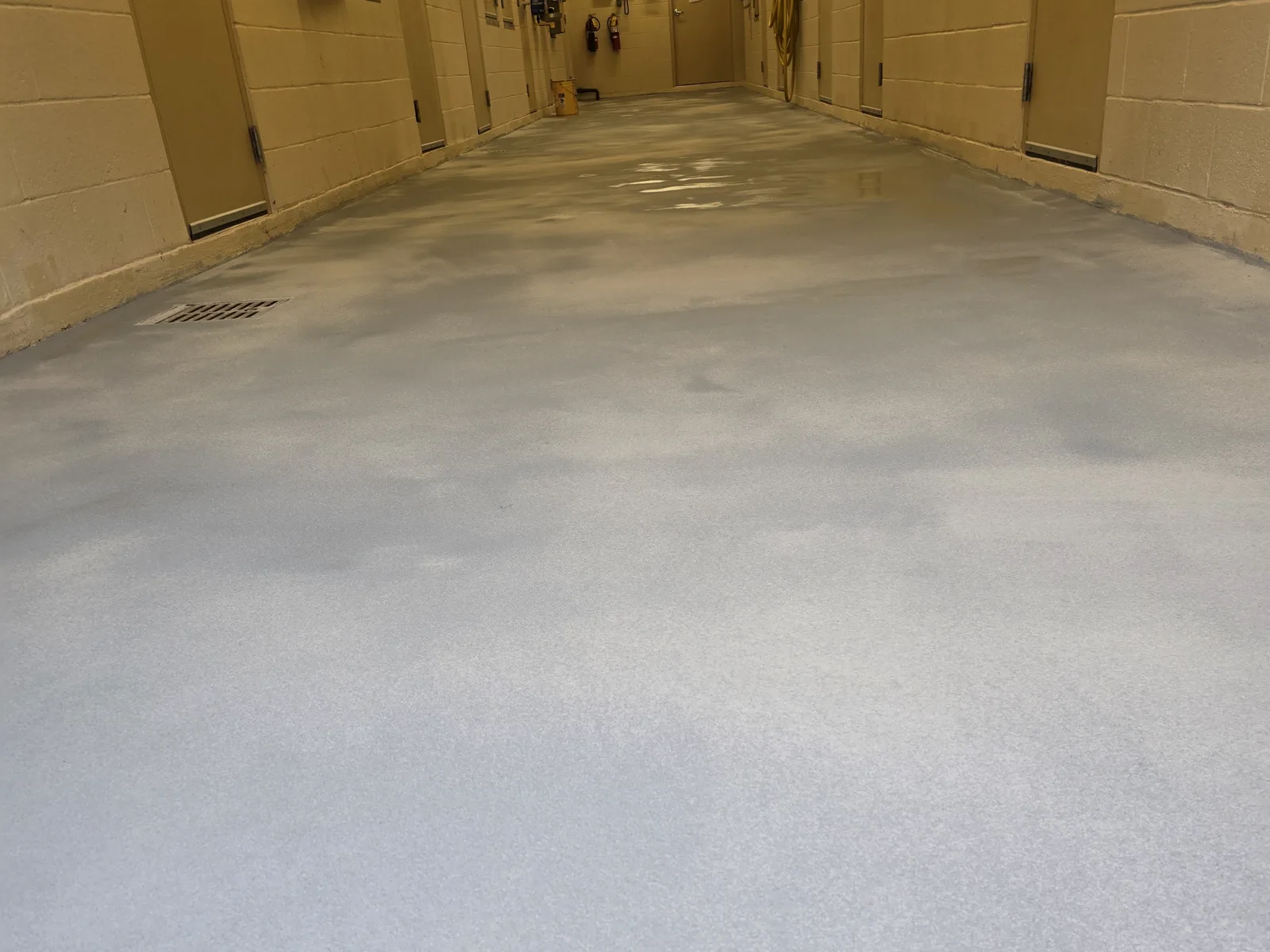 A back-of-house corridor with painted block walls and heavy doors, floored with a pale grey textured coating that turns up the wall as a coved base, with a floor grate set into it near the left wall.