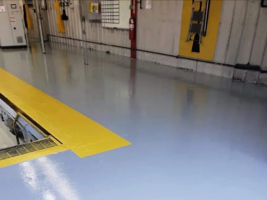 An aisle in a working facility with a pale blue-grey coated floor, yellow safety striping marking the edge of a grated service pit, machinery and a fire extinguisher along the wall behind.