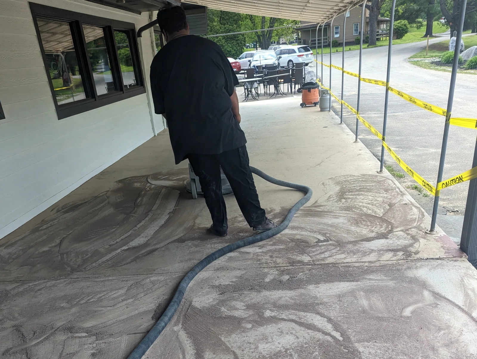 A worker seen from behind guiding a walk-behind grinder across a dusty concrete slab under a covered walkway, with a grey dust-extraction hose lying across the floor.
