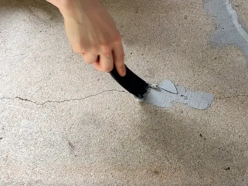 A hand using a brush to work grey repair material into a fine crack running across a rough concrete slab, with an earlier repaired area visible nearby.