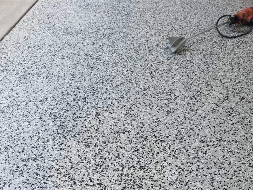 Looking down on a dense black, white and grey decorative broadcast covering a floor, with no sheen on the surface and a mixing paddle resting at the edge of the frame.