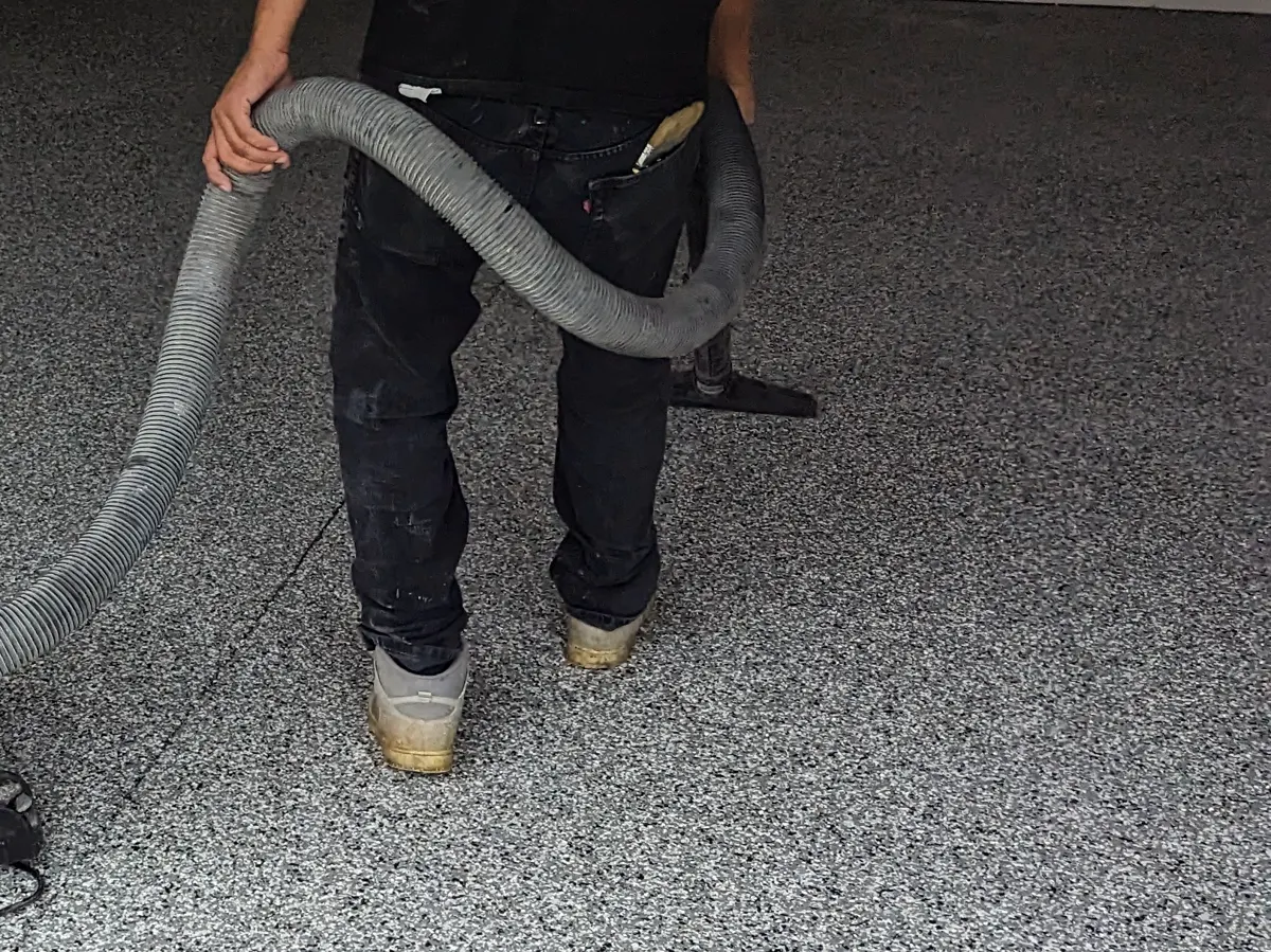 An installer seen from the waist down drawing a vacuum wand across a dense grey and black flake floor to lift loose material from the surface.