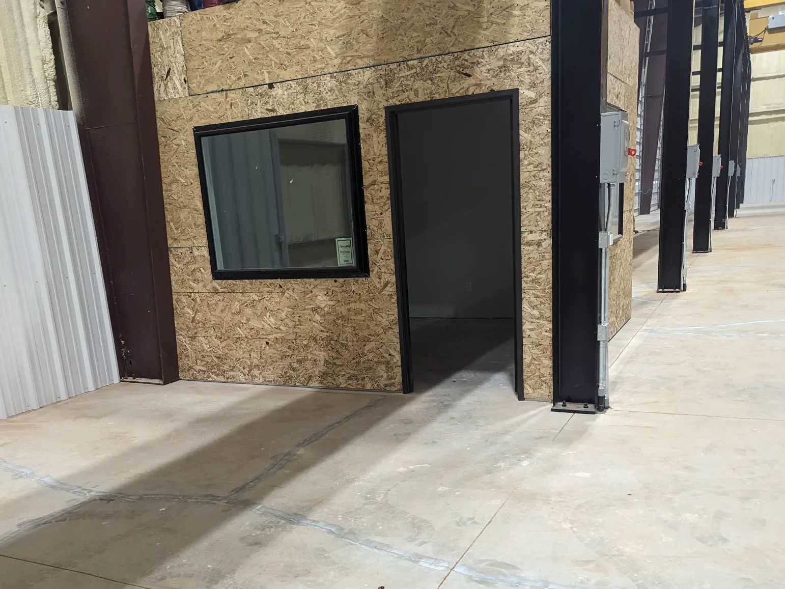 A bare concrete slab in a steel-framed industrial building with saw-cut joints, alongside a timber-clad internal office structure with cable reels stored on top and electrical disconnects mounted on the columns.