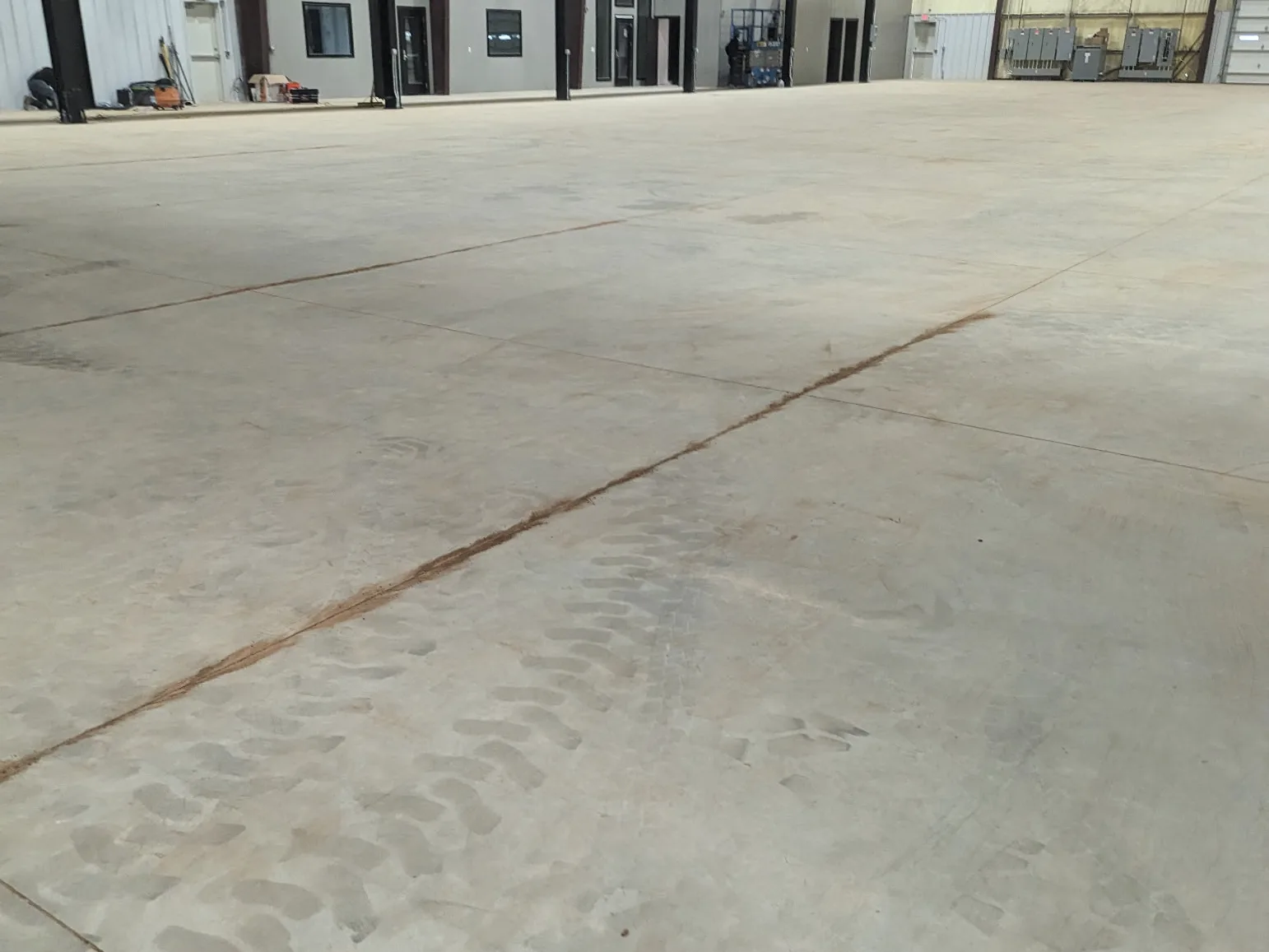 A large bare concrete slab inside a steel-framed industrial building, marked with equipment tyre tracks and crossed by saw-cut joints, with electrical switchgear and an internal plant room along the far wall.