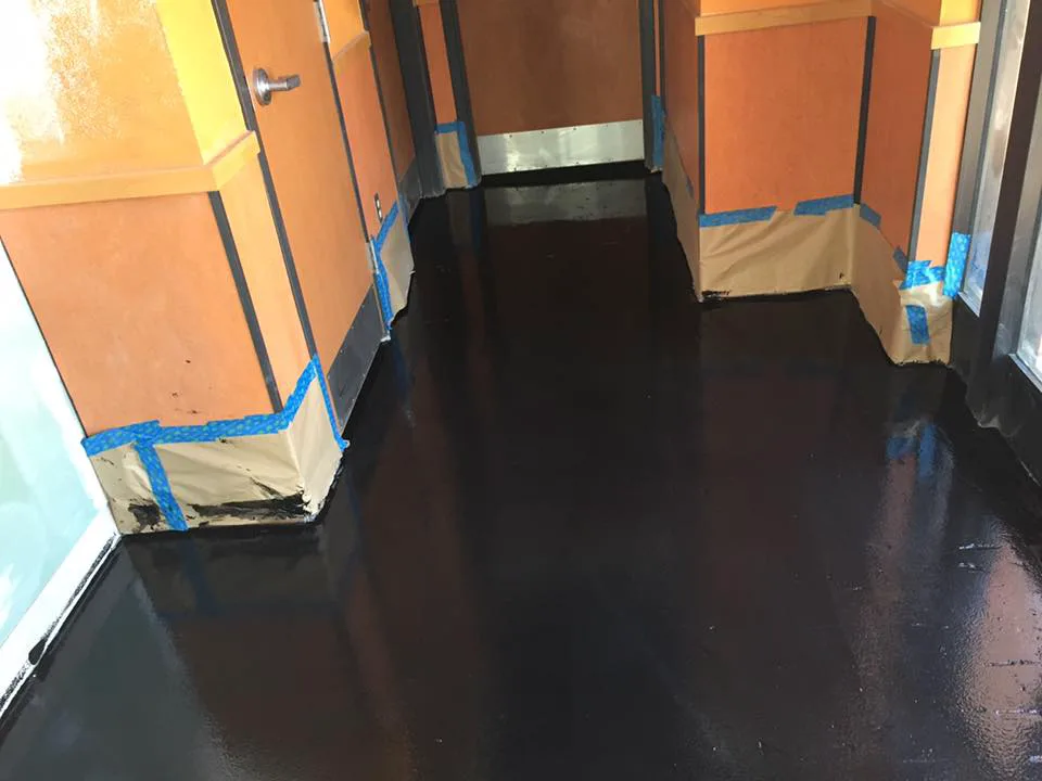 A corridor during a coating installation, with wall bases masked in paper and tape and a dark wet coating covering the floor between the doorways.