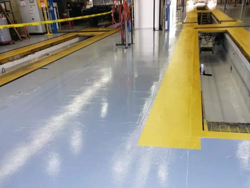 An aisle in a vehicle service facility with a pale blue-grey coated floor running between two long service pits edged in yellow, with hose reels, cabinets and equipment along the wall behind.