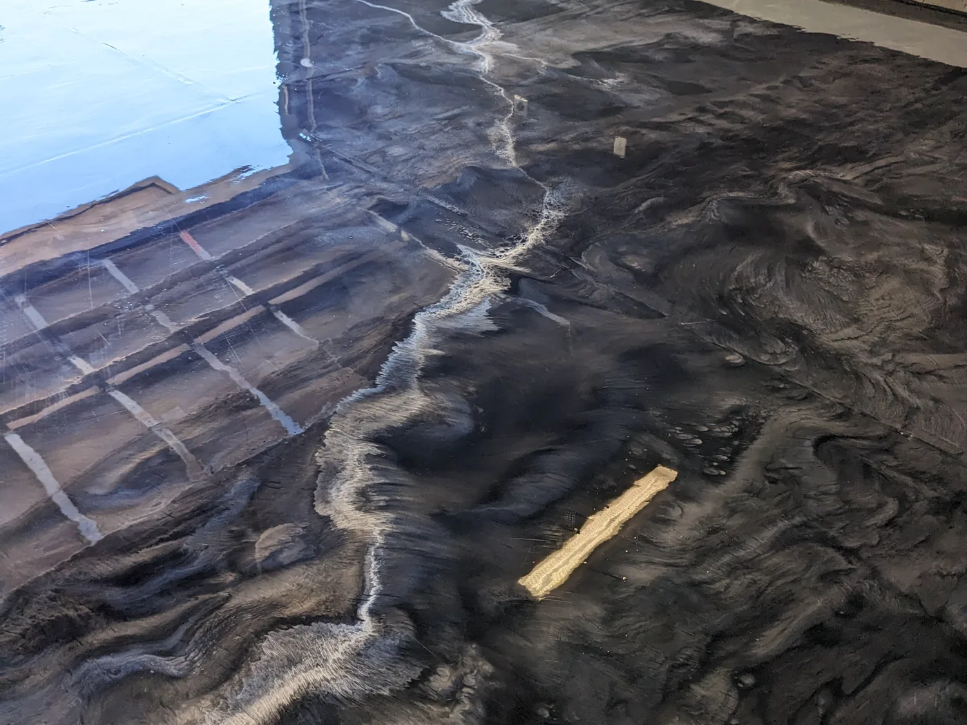 A black and silver metallic coat being worked across a floor, with pale veining running through the dark base and a spreading tool resting on the wet surface.