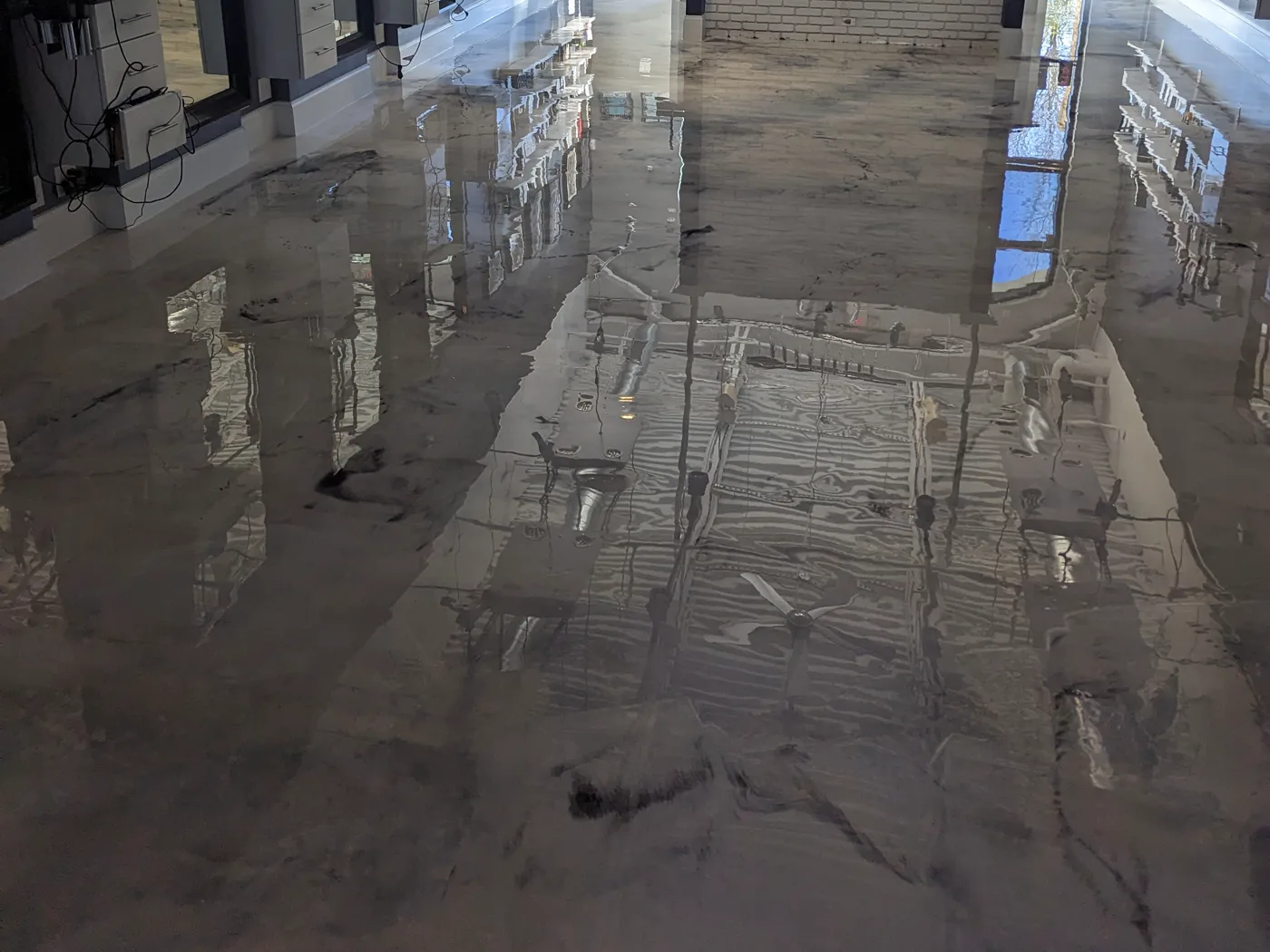 A grey and white marbled metallic floor running the length of a commercial interior, its mirror finish reflecting fitted furniture, ceiling lights and daylight from the windows.