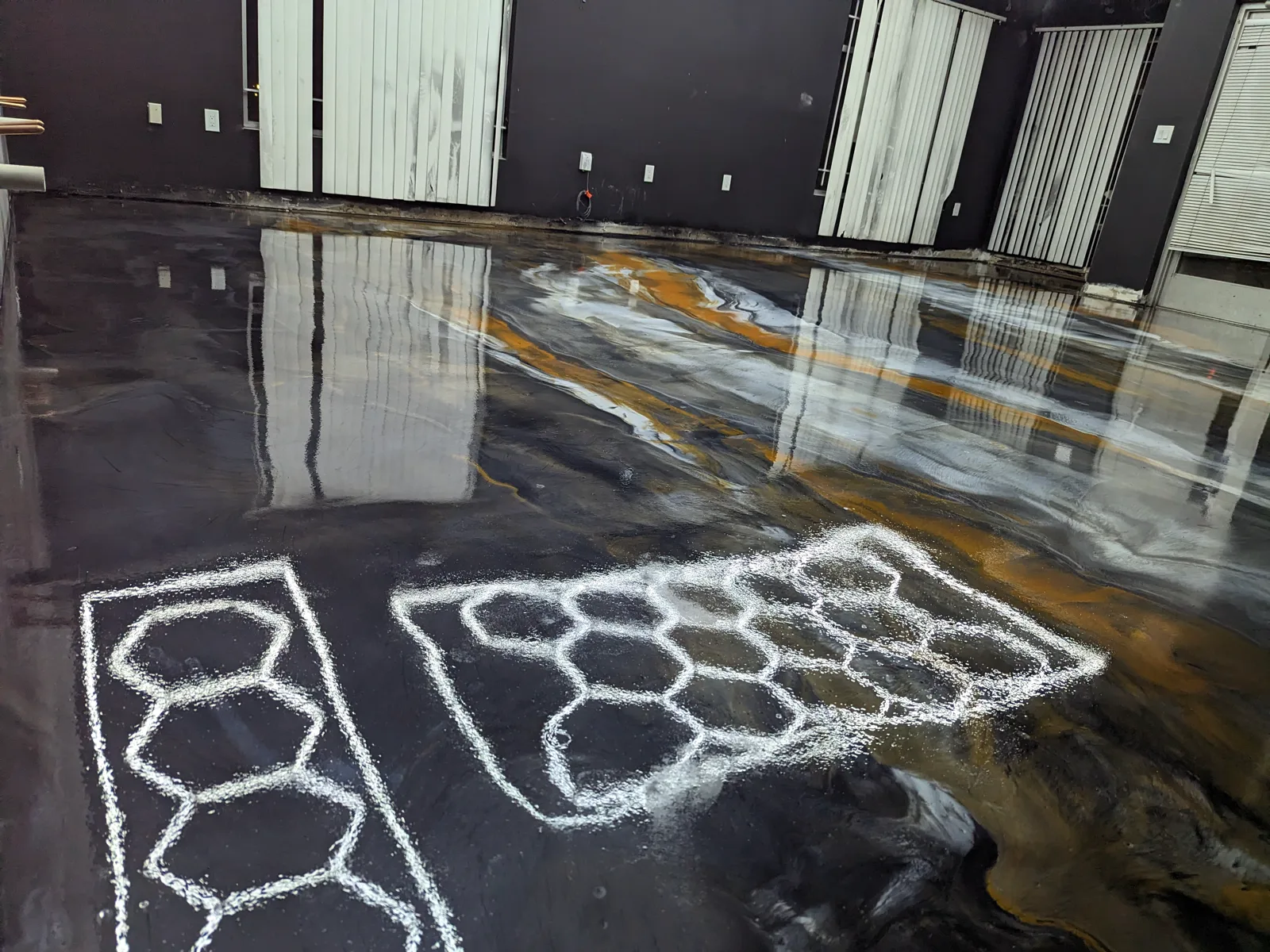 A black, grey and amber metallic floor still wet and fully reflective, with veining running across it and the hexagonal ceiling light mirrored in the surface.